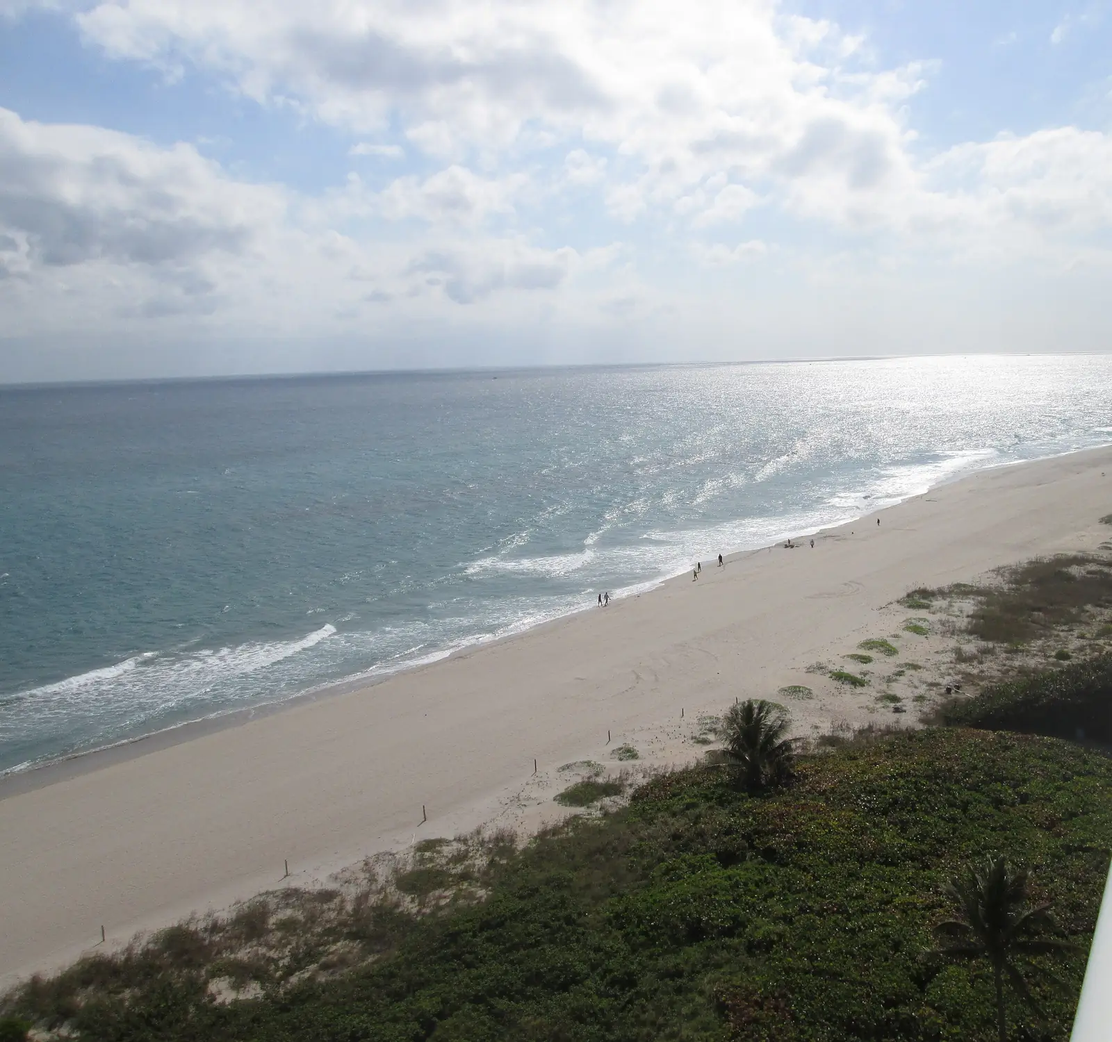 High view south along a long wide beach with walkers at the waterline, sea-oat dunes and two palms, glittering ocean under broken cloud; the curved balcony railing and the building's facade fill the right edge.