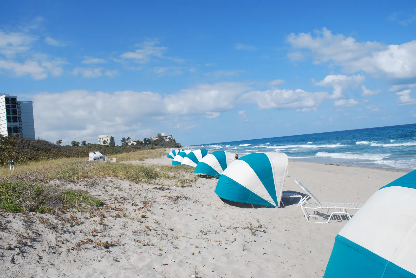 Sunny beach scene: dune grasses and sand in the foreground, a row of teal-and-white striped beach cabanas and loungers along the water, surf breaking at right, a condominium tower at far left and puffy clouds.