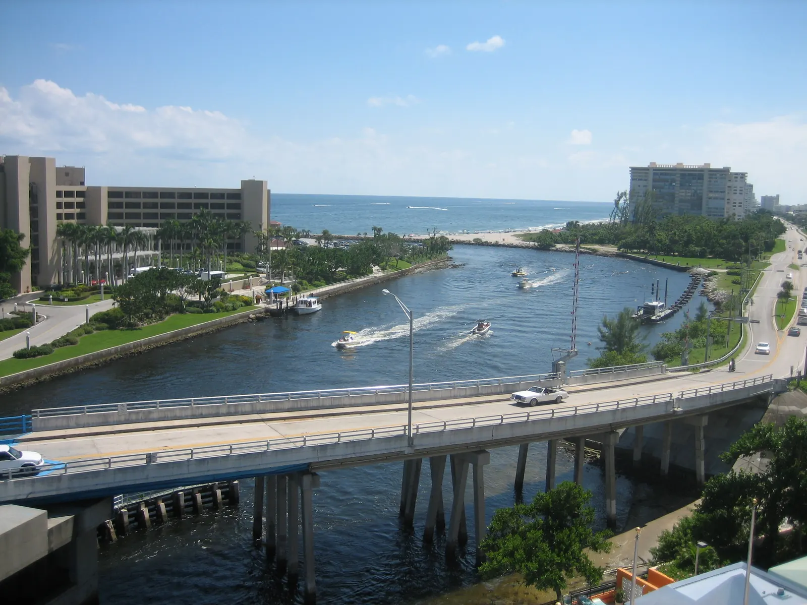 Elevated view down the Boca Raton Inlet channel to the ocean: a concrete road bridge with a white car crossing in the foreground, boats with wakes in the channel, landscaped banks, a large hotel building at left and a condominium tower at right.