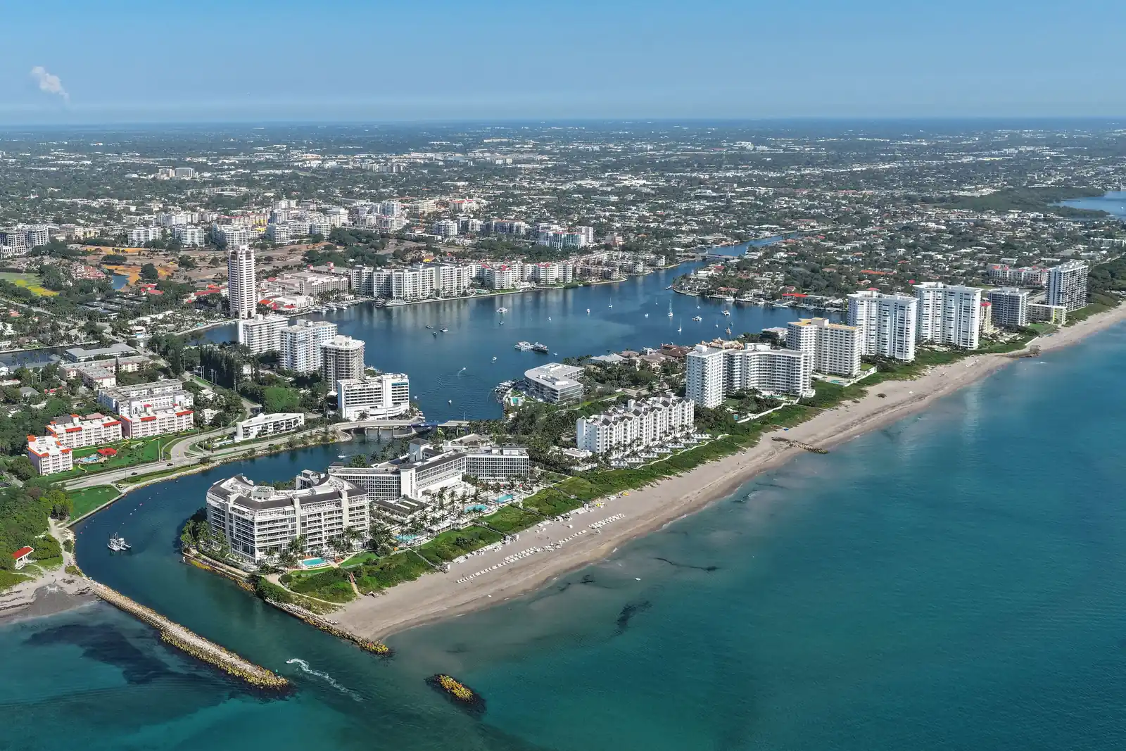 High aerial view north over the Boca Raton Inlet: two rock jetties reach into turquoise water at lower left, a long sand beach runs up the right side, white mid-rise condominium towers line the barrier island, Lake Boca Raton opens in the middle with boats, and the low city spreads to the horizon under a pale blue sky.