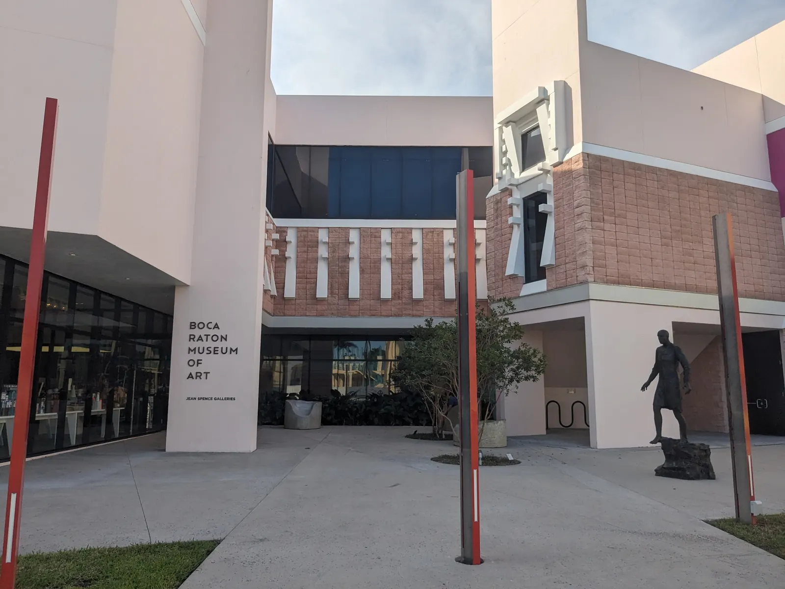 Entrance courtyard of a pink and white modernist building; a white pillar lettered "BOCA RATON MUSEUM OF ART", glass gallery walls, a bronze walking-figure sculpture on a rock at right and slender red sculpture poles in the foreground.