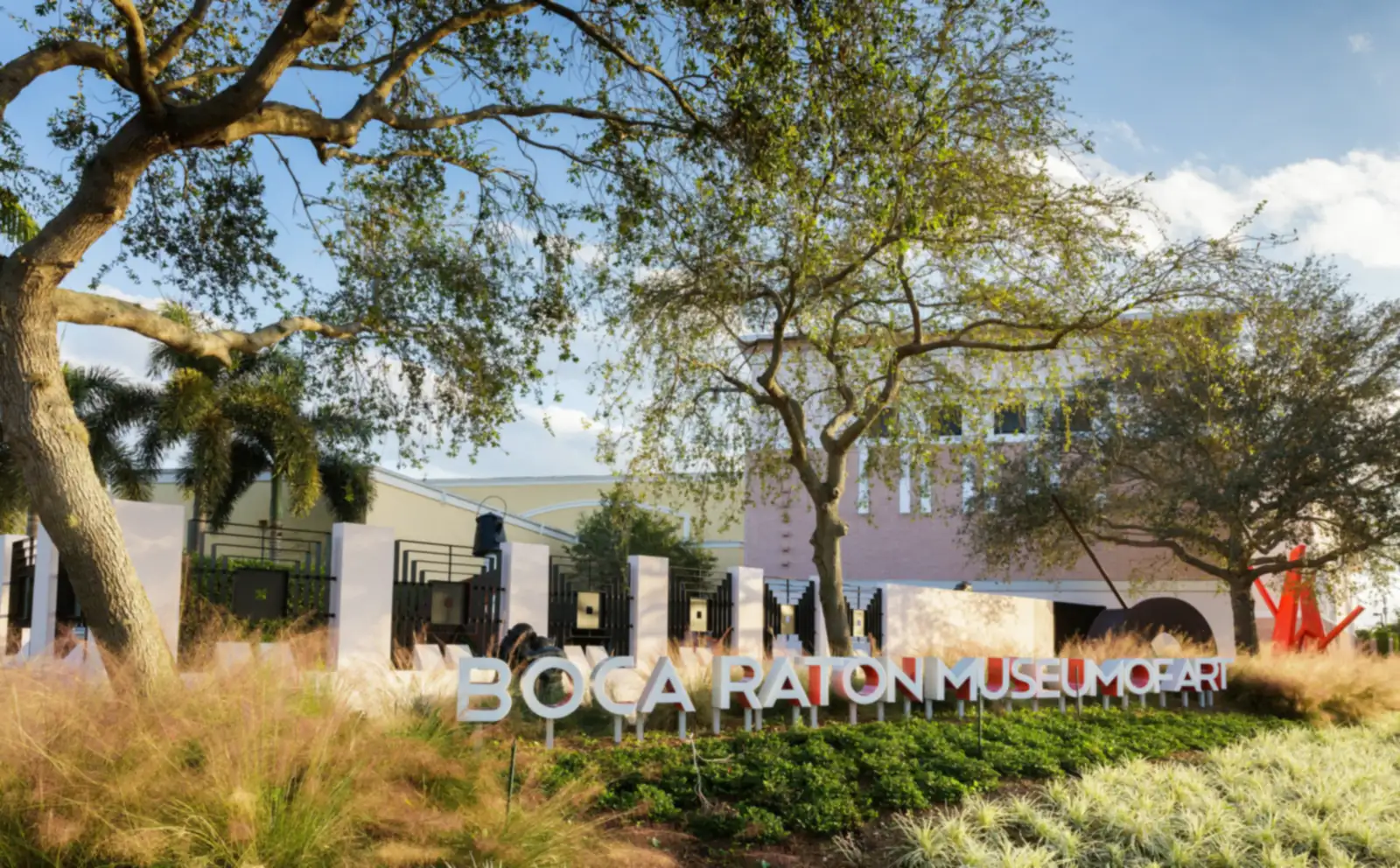 Large white three-dimensional letters "BOCA RATON MUSEUM OF ART" set in ornamental grasses and ground cover under spreading live oaks, a red abstract sculpture and the museum building behind.