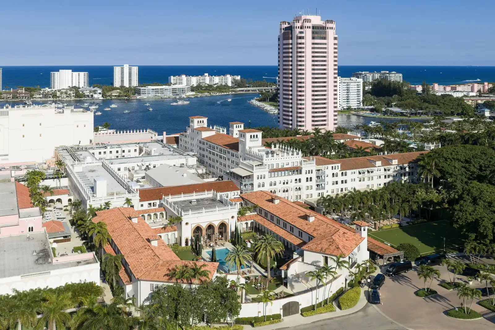 Elevated view over the resort: white Mediterranean-revival buildings with red barrel-tile roofs and an arched courtyard with a pool in the foreground, a tall pink tower at upper right, Lake Boca Raton and boats behind, ocean-front condominium towers and the Atlantic on the horizon.