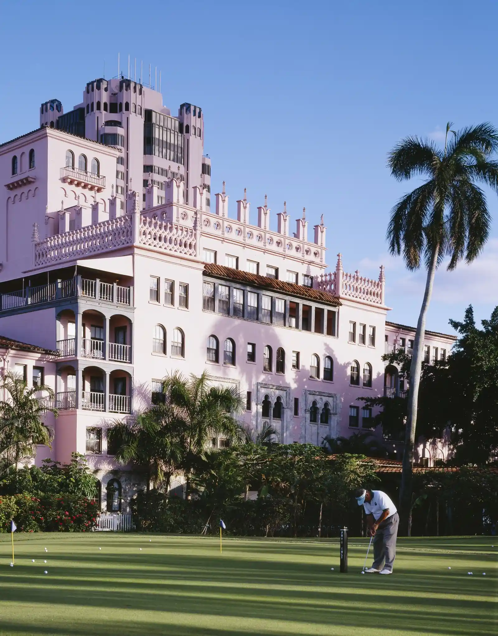 Portrait-format photo of the pink Mediterranean-revival hotel with crenellated towers and loggias above palm trees, and a golfer putting on a manicured green in the foreground.