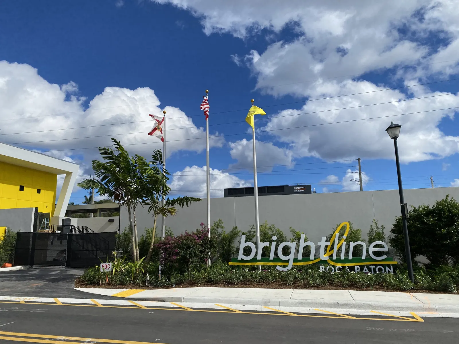 A landscaped street-front sign reading "brightline BOCA RATON" below three flagpoles (Florida, US, and a yellow flag), a yellow-and-white station building at left and a grey garage wall behind, big white cumulus clouds in a blue sky; overhead wires cross the sky.