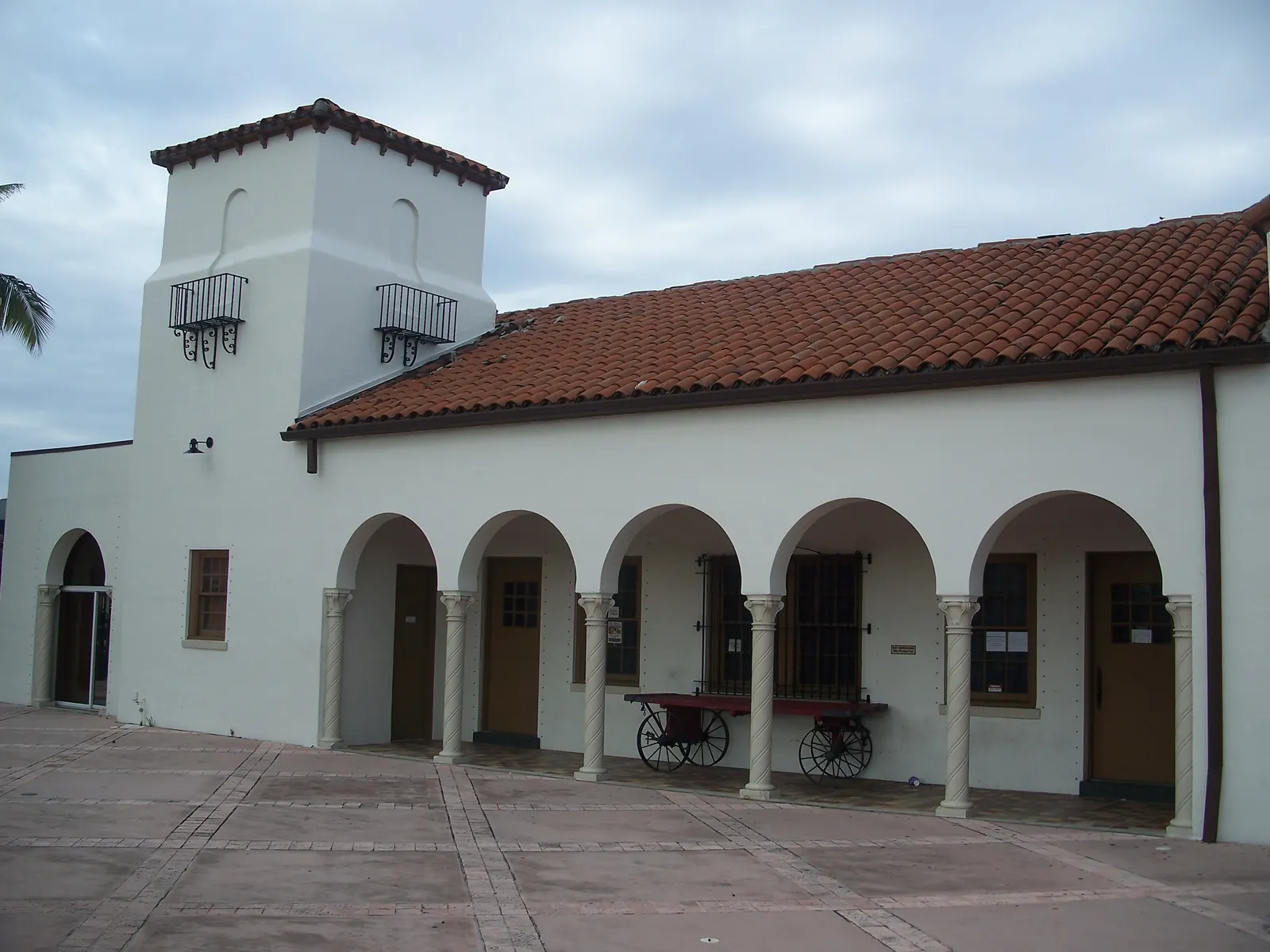 White stucco railway station with a square tower, wrought-iron balconettes, a red barrel-tile roof and an arched loggia with twisted columns; a red antique baggage cart under the arches and a paver platform in front, cloudy sky.
