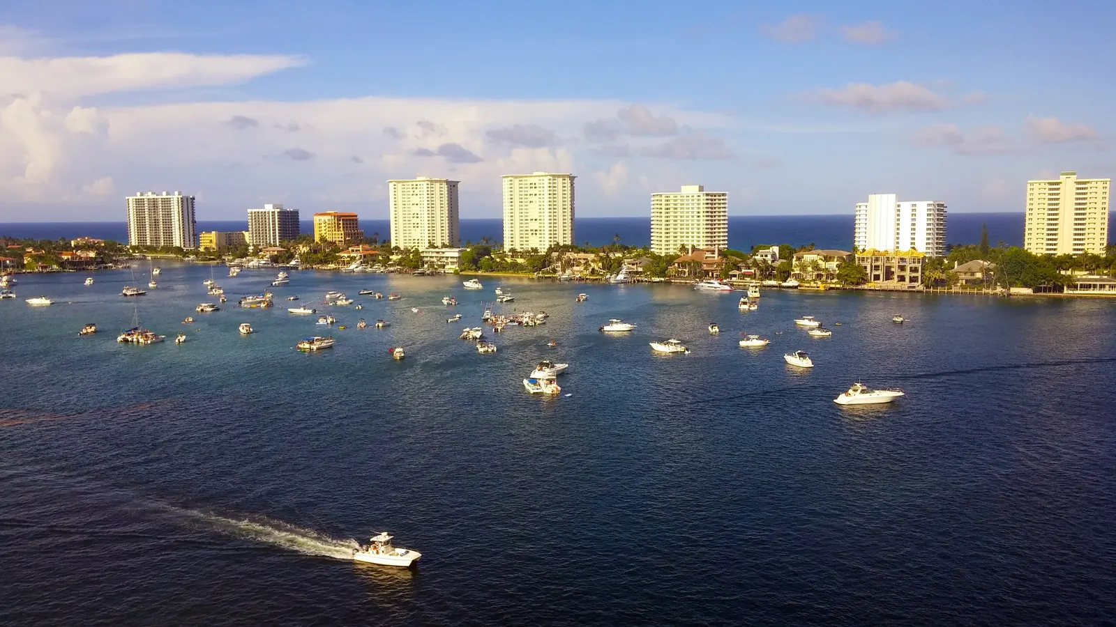 Late-afternoon light on dark-blue water dotted with dozens of anchored boats, a speedboat trailing a wake in the foreground, and a row of white and cream condominium towers along the barrier island under scattered clouds.