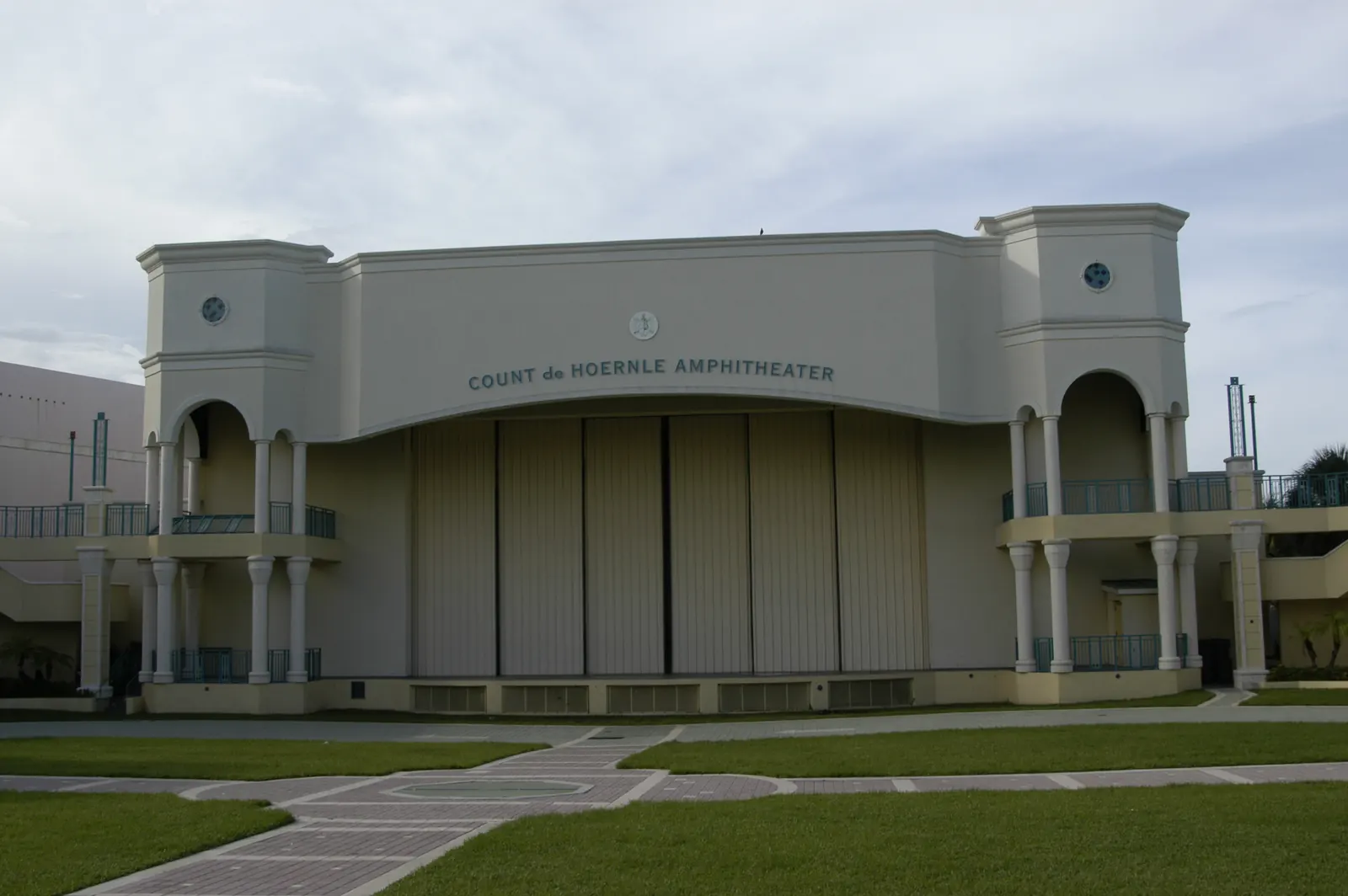 Cream stucco stage house with arched side loggias and the lettering "COUNT de HOERNLE AMPHITHEATER" across the top, closed stage doors, a lawn with paver walks in front, overcast sky.