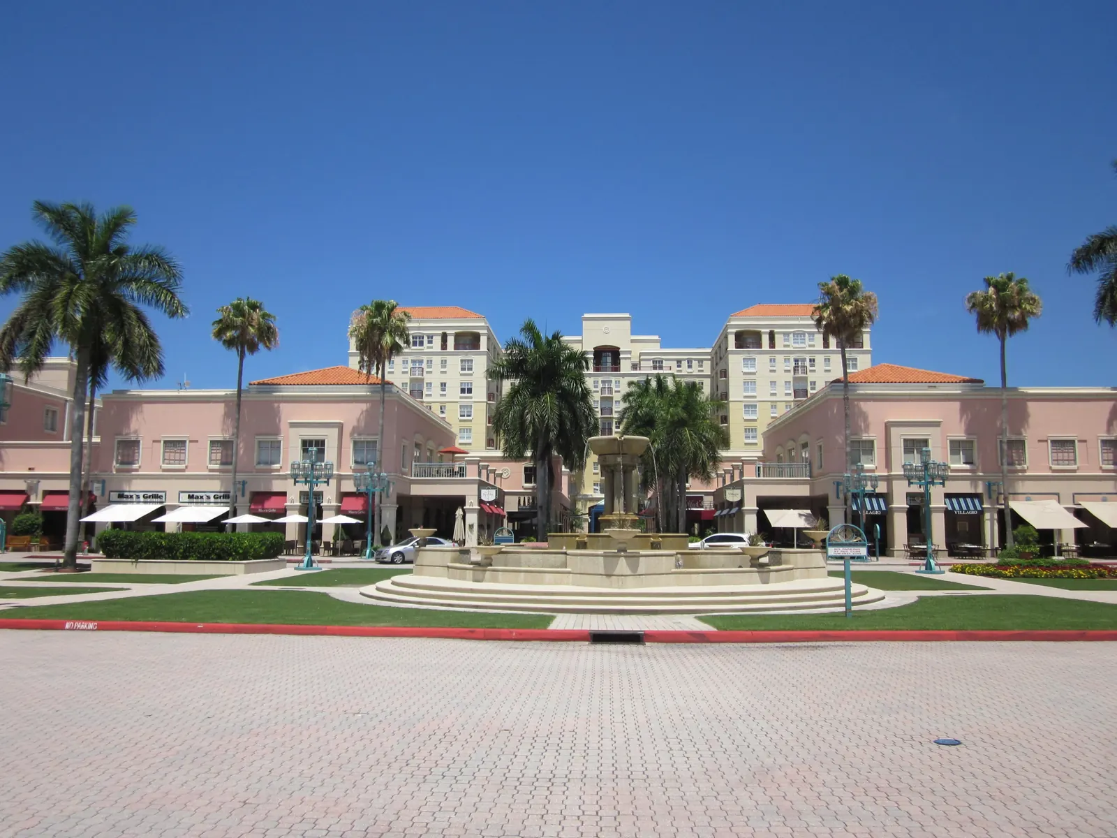 Wide sunny view across a paver plaza and lawn to a tiered stone fountain, framed by pink two-storey arcaded shops with awnings and umbrellas, cream mid-rise residences behind and royal palms against a deep blue sky.