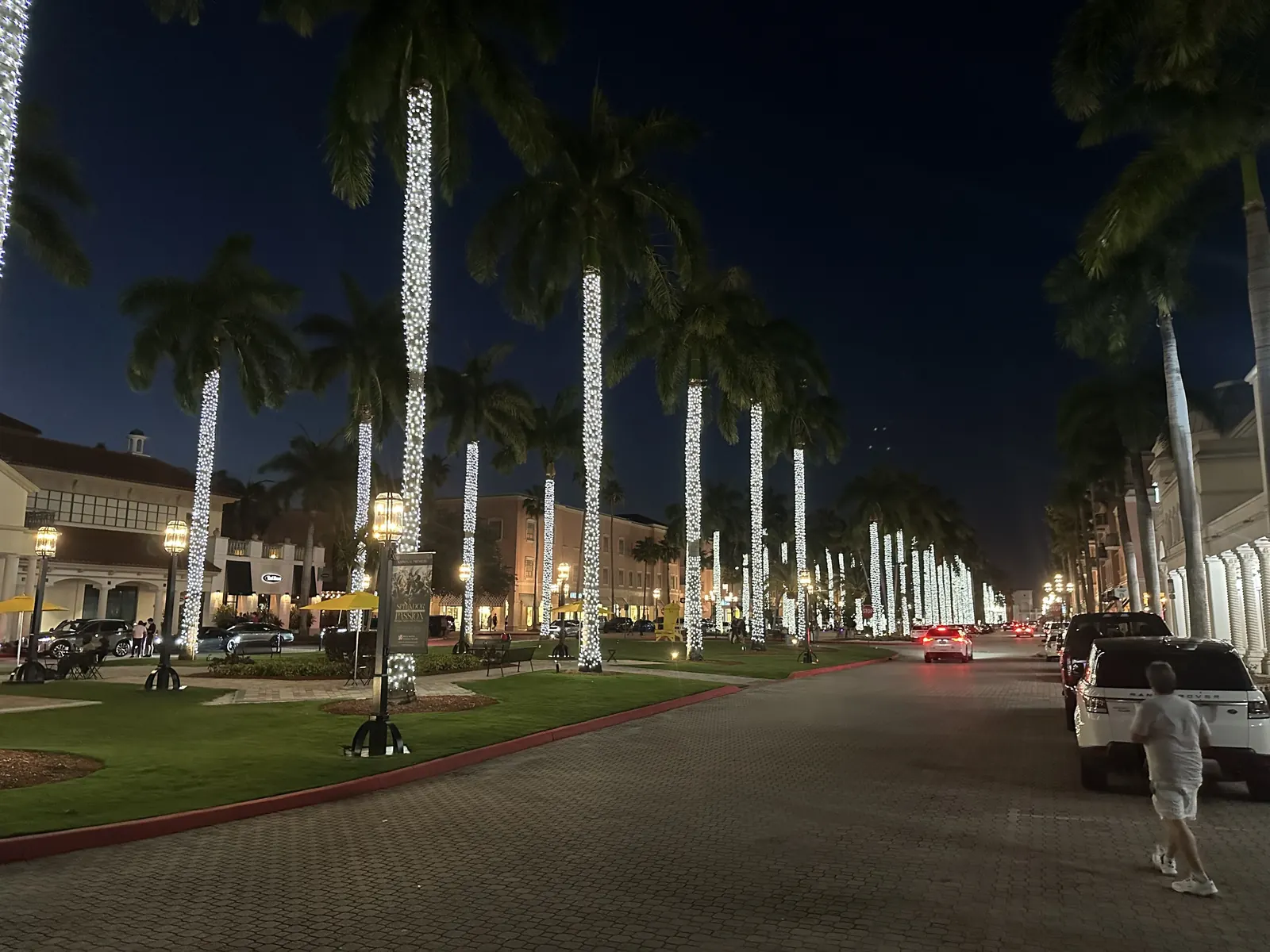 Night view down the Mizner Park boulevard: royal palm trunks wrapped in white lights recede in two rows, lantern street lamps, a lawn median, parked cars along the right, shop arcades on both sides and a man walking at right edge.