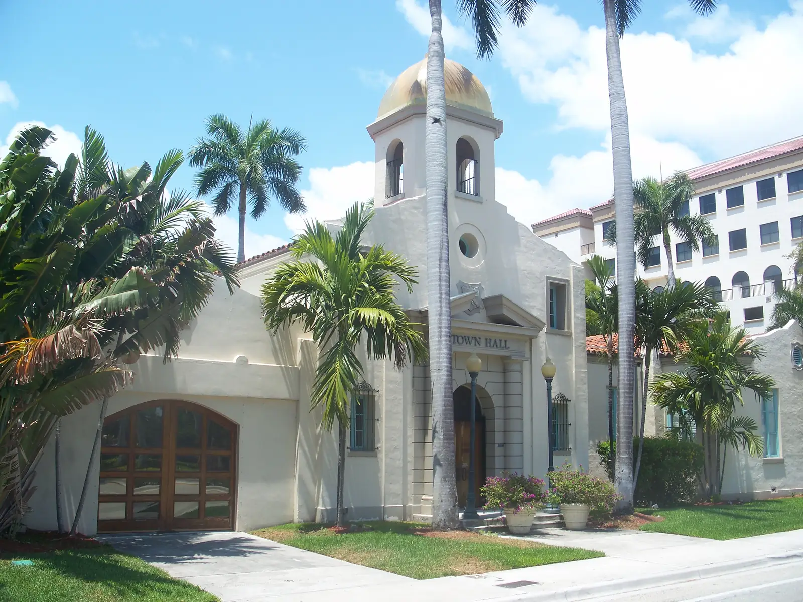 Cream stucco Mediterranean-revival building with a gold-domed bell tower, "TOWN HALL" lettering over an arched entry, a large arched wooden carriage door at left, palms and potted flowers, and a modern mid-rise building behind at right.