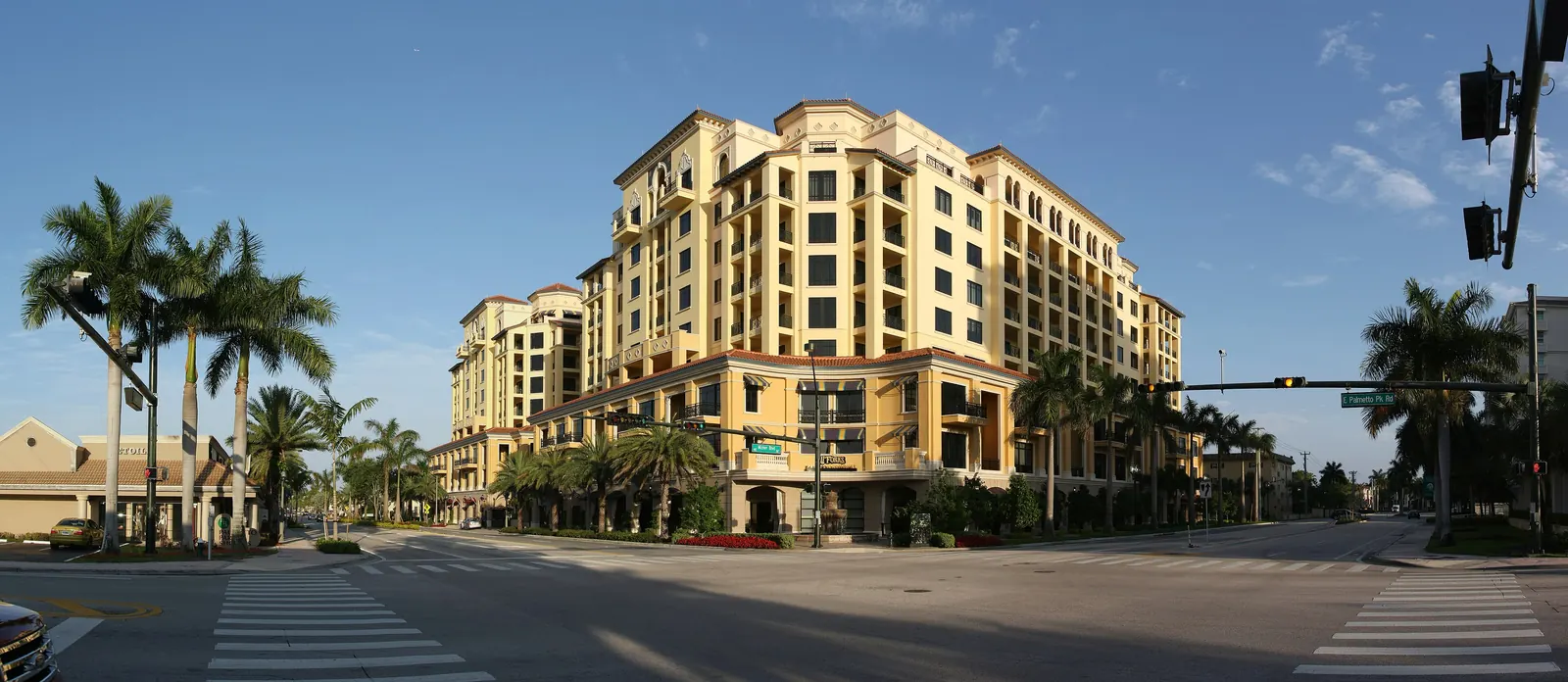 Wide stitched panorama of an empty intersection with crosswalks, a tall cream-and-ochre Mediterranean-style mixed-use building with arcades and balconies in late sun, royal palms at left and traffic signals at right.
