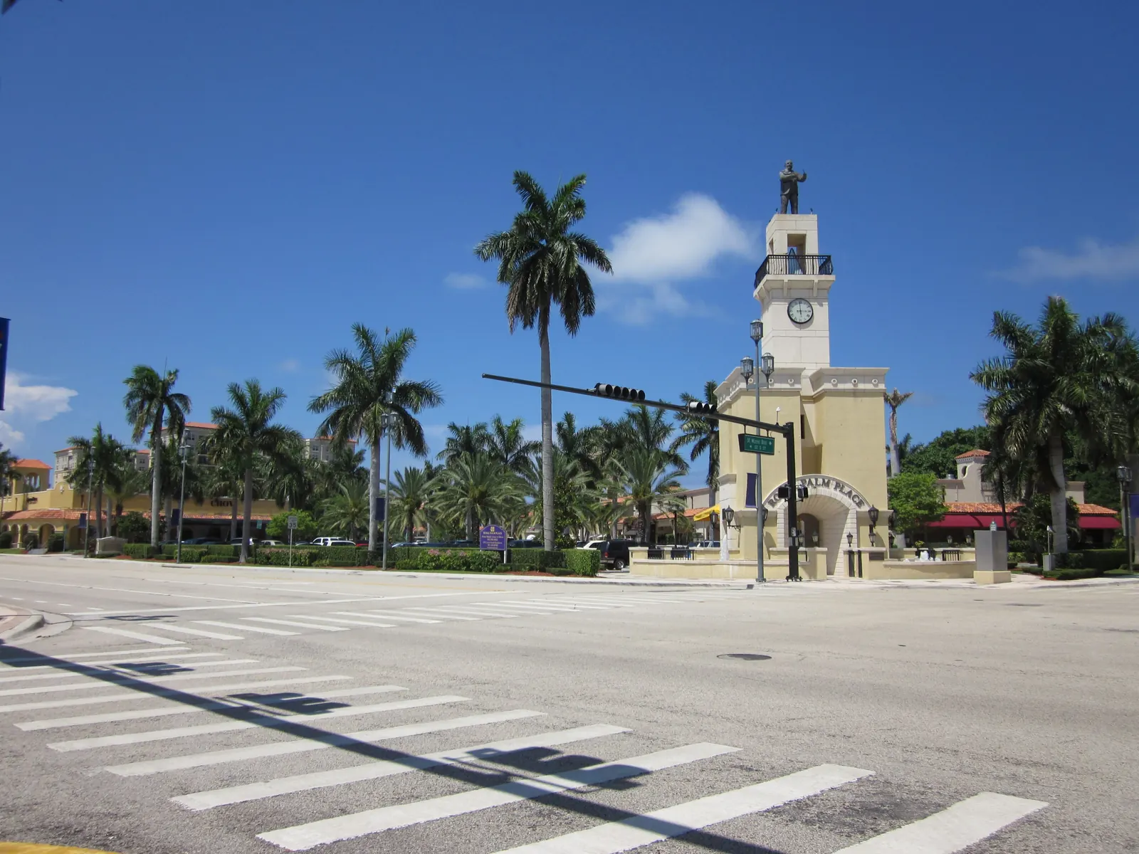 A wide sunlit intersection with a zebra crosswalk in front of a cream clock tower topped by a statue, a "Royal Palm Place" arch and sign, a traffic-signal mast arm, royal palms and low red-roofed shops under a deep blue sky.