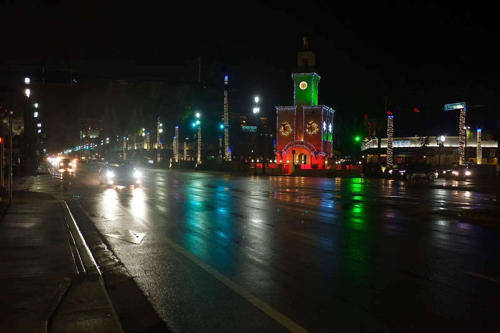 Night on a rain-wet Federal Highway: the Royal Palm Place clock tower lit red and green with holiday wreaths, car headlights and traffic signals reflecting on the pavement, light-wrapped palms along the median.